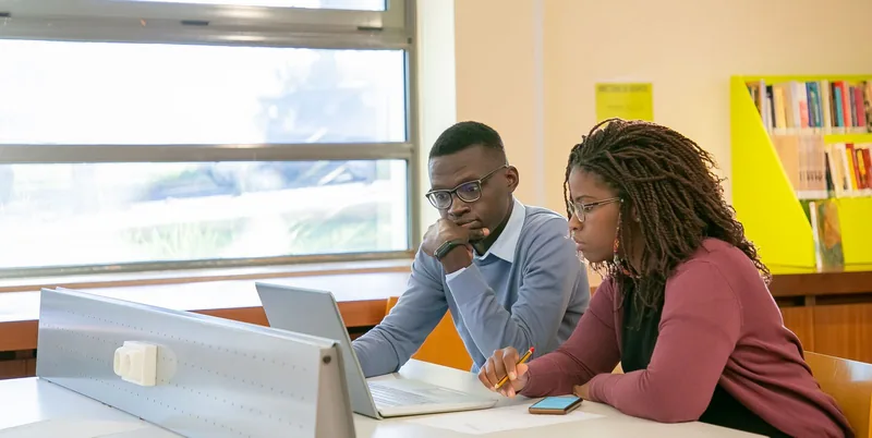 Two university students study together at a library desk with a laptop and notebook.